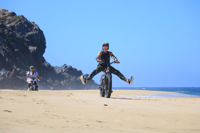 Cyclist in helmet and pads rides a fat-tire bike on a sandy beach near rocky cliffs, legs kicked out in a playful split while another rider follows along the ocean shoreline under a clear blue sky.