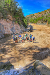 Group of off-road adventurers waving on a sandy canyon floor beside a line of UTVs, surrounded by rocky cliffs, scrubby hills and a bright blue sky with a small stream splash in the foreground.