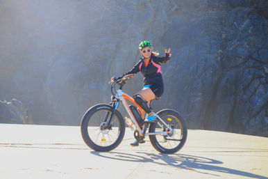 Smiling woman wearing a green helmet and sunglasses rides a fat-tire e-bike on a sunlit road, flashing a peace sign against a dramatic rocky cliff backdrop.