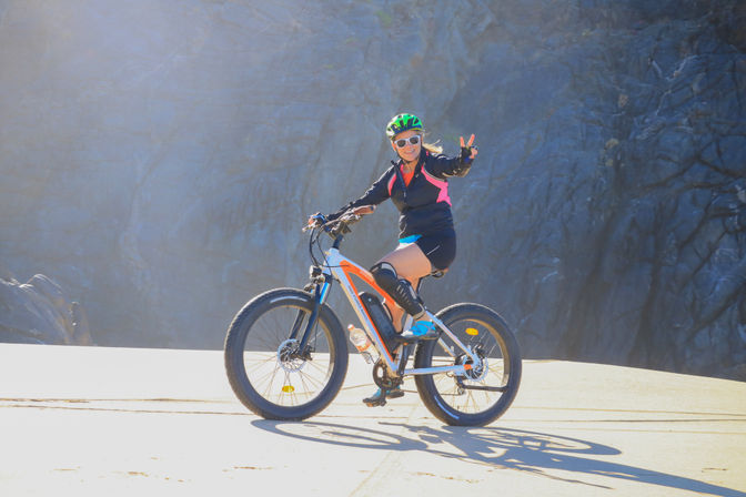 Smiling woman wearing a green helmet and sunglasses rides a fat-tire e-bike on a sunlit road, flashing a peace sign against a dramatic rocky cliff backdrop.