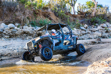 Off-road side-by-side UTV splashing through a shallow rocky creek in an arid canyon, helmeted driver steering over muddy water with rocky banks and scrub vegetation