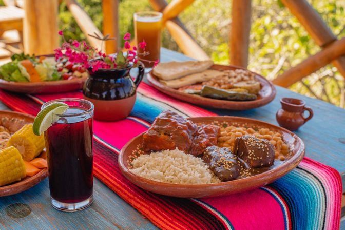 Rustic outdoor Mexican meal on a colorful serape: clay plates with rice, beans, mole-topped meat sprinkled with sesame, corn, tortillas, salad, and a glass of hibiscus agua fresca garnished with lime.