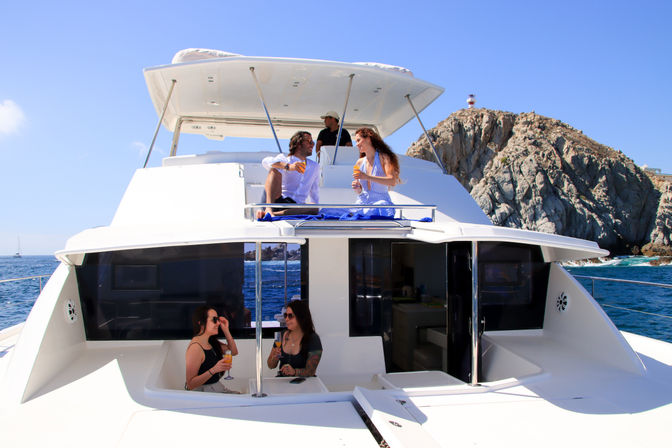 Group sipping cocktails on the deck of a white luxury yacht cruising past rugged coastal cliffs and a lighthouse with bright blue ocean and clear sky