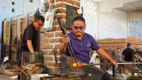 Glassblower shaping glowing orange molten glass with a long rod in a brick-walled workshop, fellow artisans and furnaces visible in the background