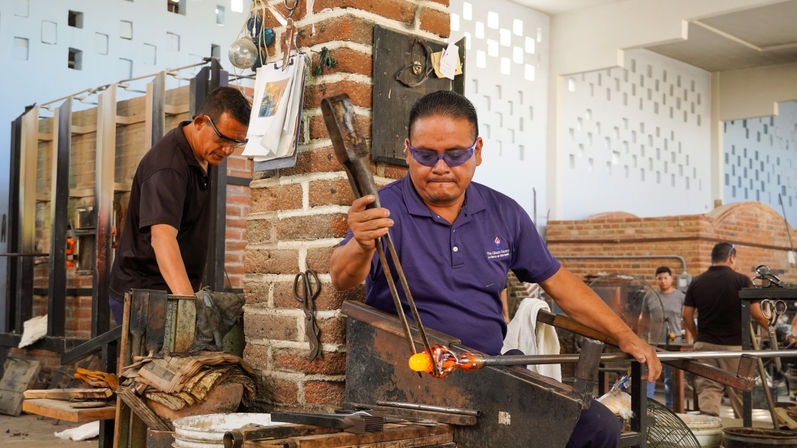 Glassblower shaping glowing orange molten glass with a long rod in a brick-walled workshop, fellow artisans and furnaces visible in the background