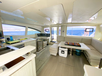 Bright modern catamaran interior showing open salon and galley with cream L-shaped seating, dining table, compact kitchen, and large windows framing blue sea and coastline with people visible on the deck.
