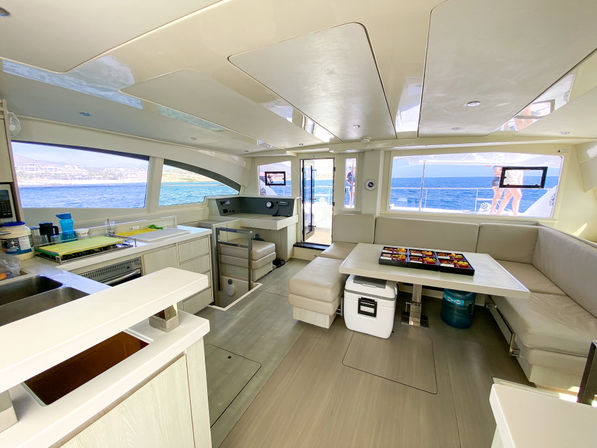 Bright modern catamaran interior showing open salon and galley with cream L-shaped seating, dining table, compact kitchen, and large windows framing blue sea and coastline with people visible on the deck.