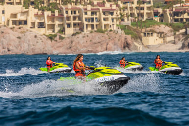 Four riders zoom on lime-green jet skis, wearing orange life jackets and splashing through choppy blue ocean water off a rocky coastline with beachfront resort buildings in the background.
