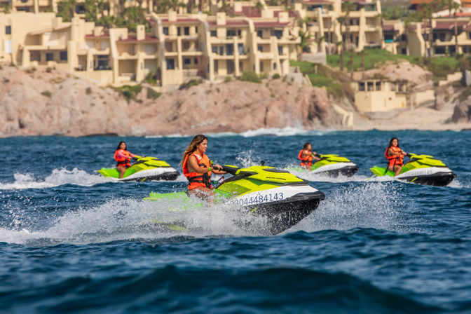 Four riders zoom on lime-green jet skis, wearing orange life jackets and splashing through choppy blue ocean water off a rocky coastline with beachfront resort buildings in the background.