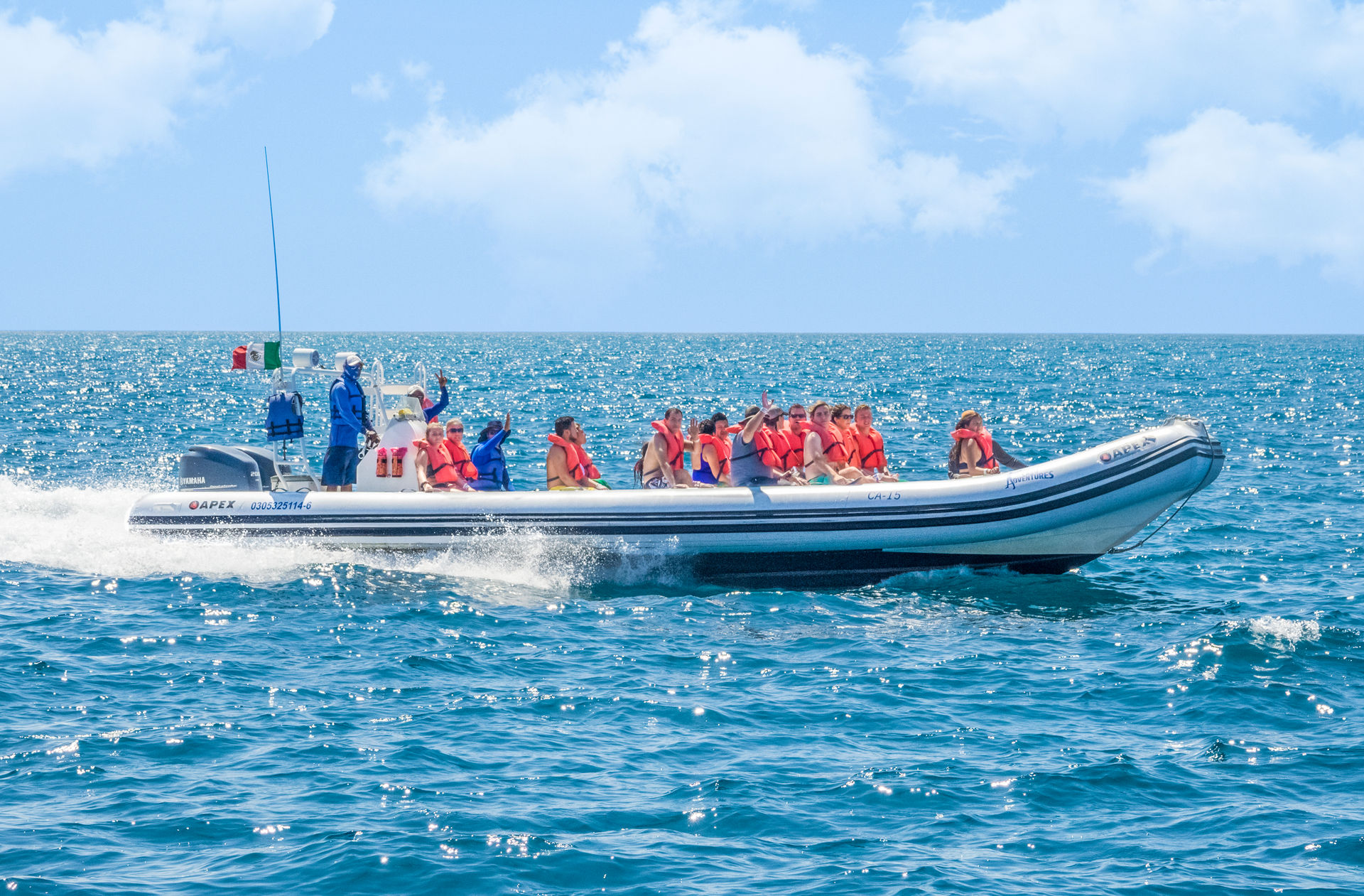 Group of passengers in orange life jackets on a white inflatable speedboat speeding across sparkling turquoise ocean on a sunny day — ocean tour vibe.