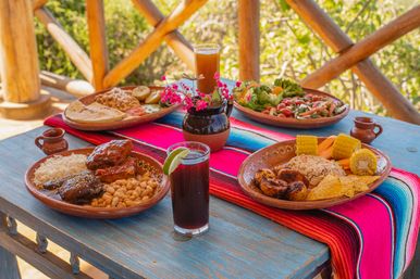 Mexican-style outdoor breakfast on a rustic blue wooden table with a colorful serape runner, clay plates of rice, beans, grilled meats and plantains, corn on the cob, salad, tortillas, a lime drink and pink flowers.