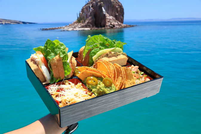 Coastal lunch box with sandwiches, tortilla chips, guacamole, jalapeños and shredded cheese held over turquoise sea with a rocky islet in the background