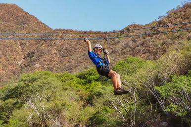 Thrilled person in helmet and harness riding a zipline over green trees and a dry mountain canyon under a clear blue sky — outdoor mountain zipline adventure.