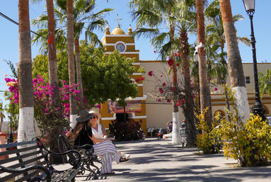 Sunny Mexican plaza with tall palm trees and pink bougainvillea, a yellow clock tower, shaded benches and two people chatting on a bench