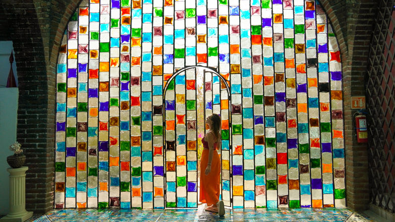 Silhouette of a person in an orange dress standing before a large arched stained-glass wall of colorful square panes in a brick archway, sunlight casting vibrant mosaic reflections on the tiled floor.