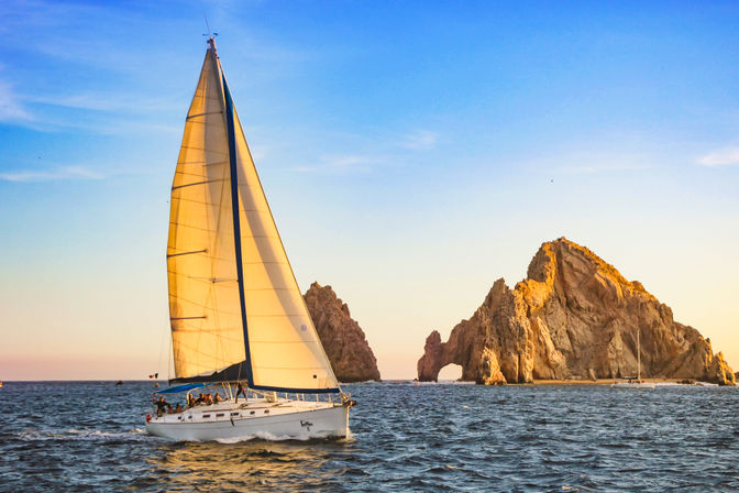 White sailboat gliding across blue sea at golden sunset with a dramatic rocky coastal arch in the background
