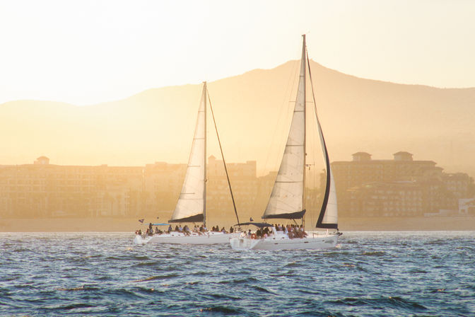 Two white sailboats with full sails gliding across sunlit blue water at golden hour, passengers on deck and hazy coastal buildings and mountains in the background.
