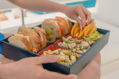 Poolside lunch bento box with sliced whole-grain sandwiches, cup of pickled jalapeños, tortilla chips and guacamole, diced chicken salad and pasta salad in compartments.
