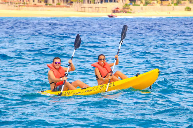 Two people in orange life jackets paddling a bright yellow tandem kayak on turquoise ocean waters with a sunny sandy beach and shoreline in the background, smiling and enjoying a coastal adventure.