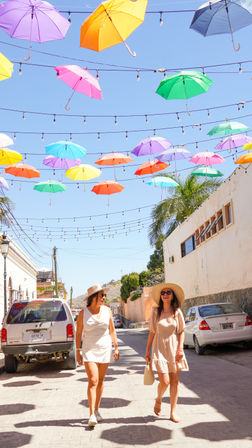 Two women in summer dresses and wide-brimmed hats strolling under a colorful suspended umbrella canopy on a sunny street with palm trees and parked cars.