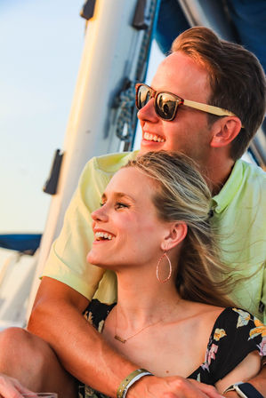 Smiling young couple embracing on a sailboat at sunset — woman with hoop earrings and floral dress, man wearing sunglasses, ocean and mast bathed in golden light