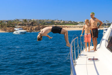 Person mid-backflip off a yacht into clear blue ocean near a sunny beachfront resort, two boys watching from the deck
