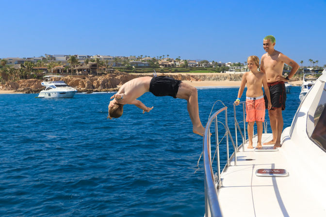 Person mid-backflip off a yacht into clear blue ocean near a sunny beachfront resort, two boys watching from the deck