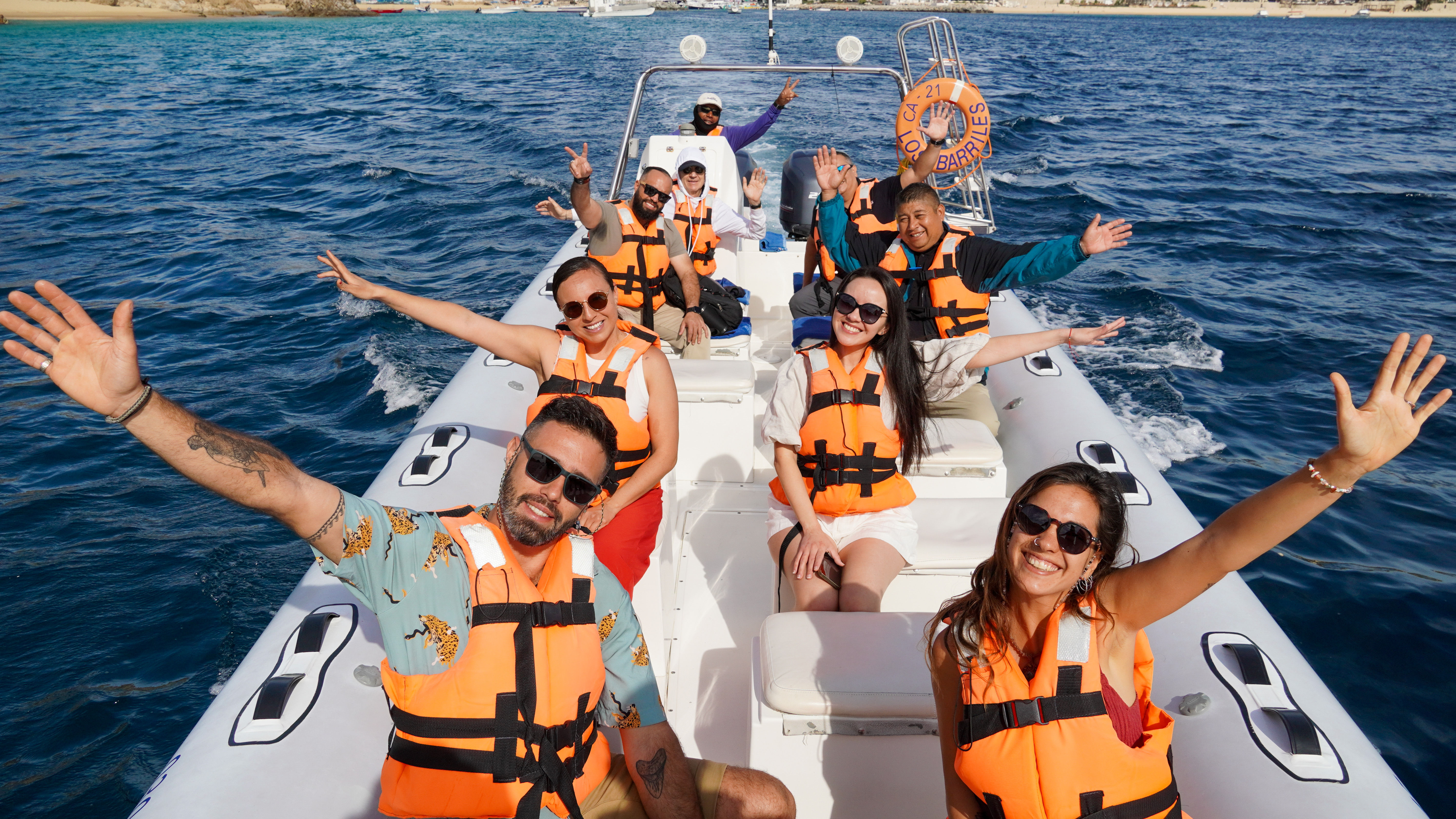 Smiling group on a white speedboat wearing orange life jackets, waving during a sunny coastal ocean tour