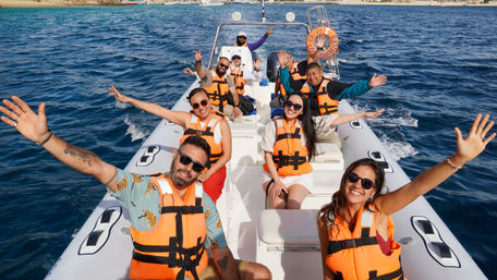 Smiling group on a white speedboat wearing orange life jackets, waving during a sunny coastal ocean tour