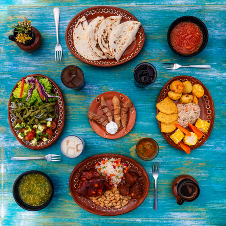 Vibrant Mexican-style spread on a turquoise wooden table: warm corn tortillas, red and green salsas, rice and beans with saucy meat, roasted potatoes, corn on the cob, chips and dip, fresh salad with nopales, and churros with cream.