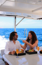 Laughing couple enjoying a luxury yacht brunch on the open blue ocean — woman feeds man while boxed sandwiches and mimosas sit on a glossy table with stainless-steel rail and sea horizon in the background.