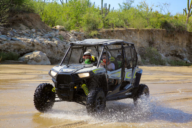 Thrilling off-road side-by-side UTV splashing through a shallow desert riverbed, riders in helmets and goggles, sand and rock canyon with cacti in the background.