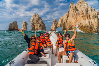 Smiling group in orange life jackets on an inflatable boat, cheering near dramatic coastal sea arch and rocky cliffs over turquoise ocean under a blue sky