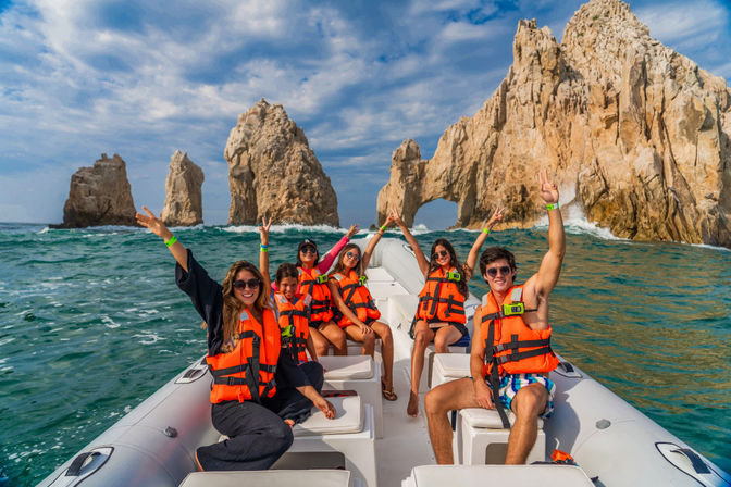 Six people in orange life jackets cheering on a small boat beside the rock arch of El Arco at Land's End, Cabo San Lucas, with turquoise water and a dramatic sky.