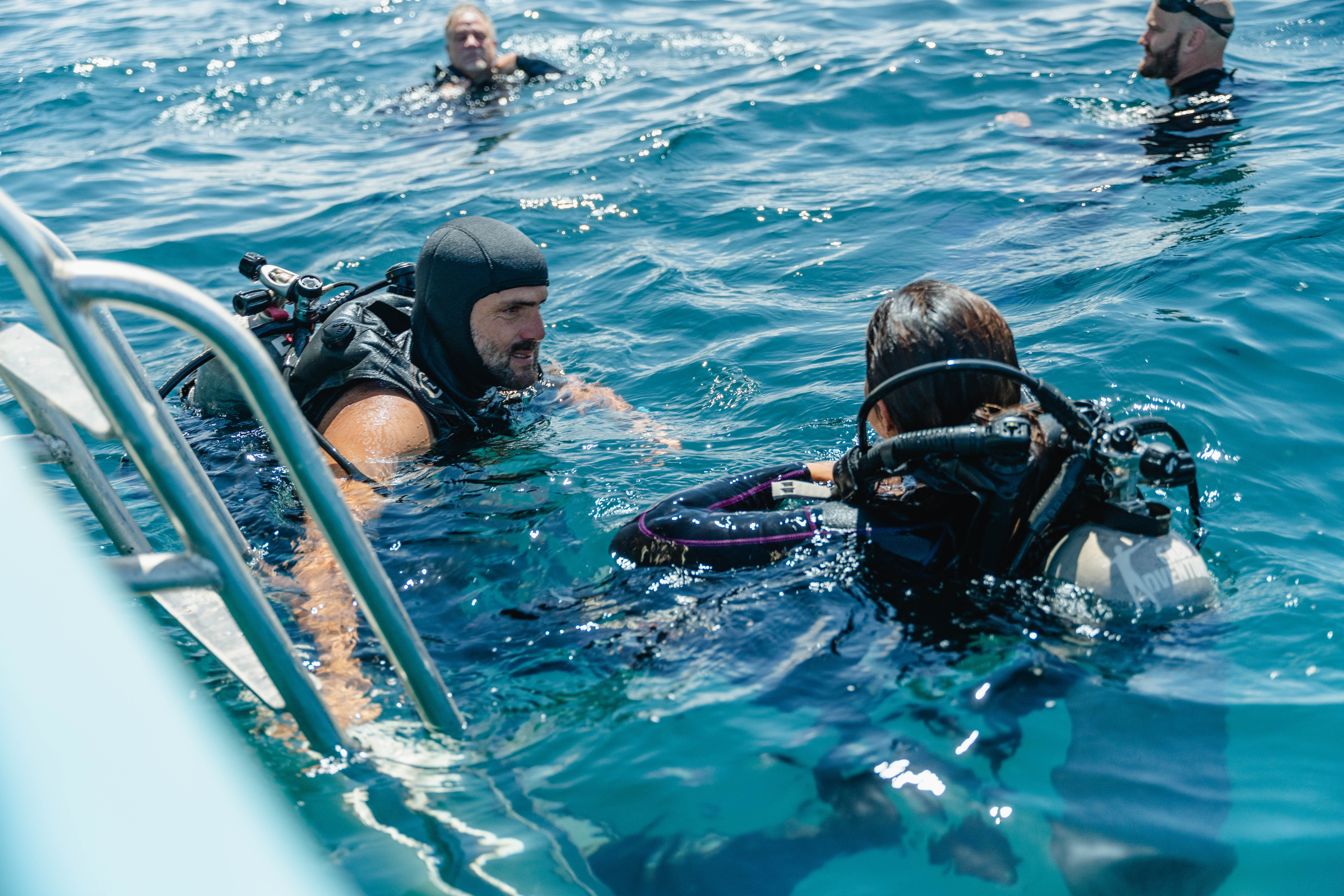 Scuba divers in sunlit clear blue open sea near a boat ladder, two checking gear and chatting while others float nearby.