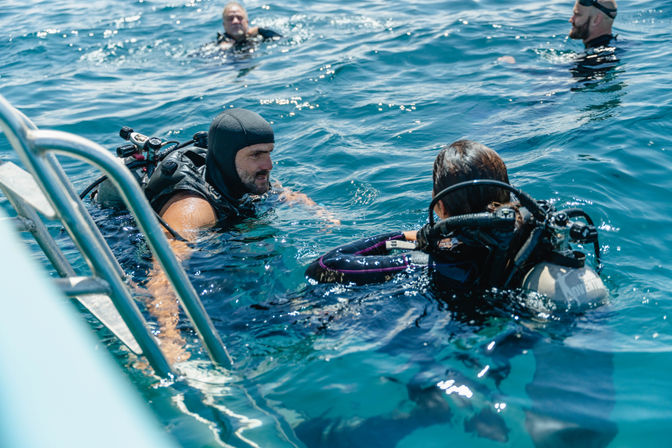 Scuba divers in sunlit clear blue open sea near a boat ladder, two checking gear and chatting while others float nearby.