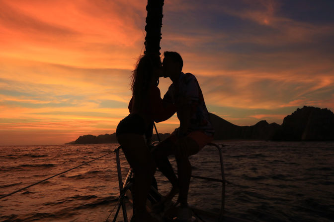 Romantic silhouette of a couple kissing on the bow of a sailboat during a vivid orange-pink sunset over the ocean with rocky coastline in the background.