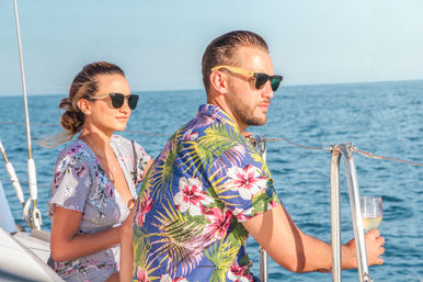 Couple in sunglasses relaxing on a sailboat in the blue open sea, man in a tropical shirt sipping white wine on a sunny sailing vacation.