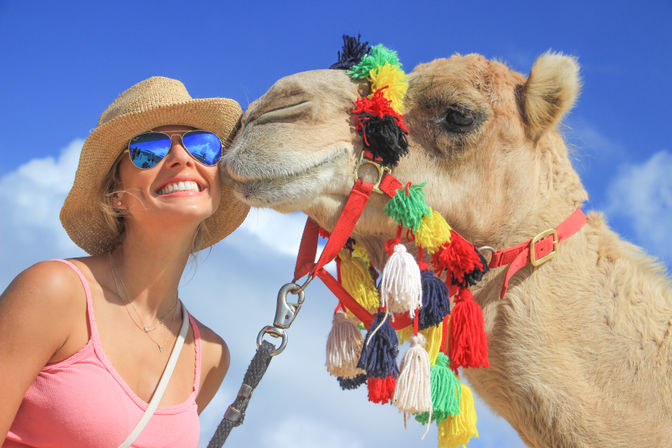 Smiling tourist in a straw hat and mirrored sunglasses posing with a camel decorated in colorful tassels against a bright blue sky.