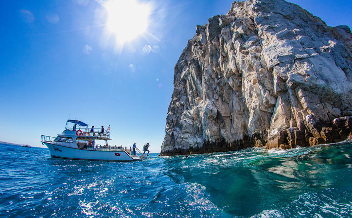 Tour boat with scuba divers leaping into turquoise ocean beside a towering rocky cliff under a bright sun and clear blue sky