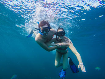 Two snorkelers wearing masks and blue fins swimming together in clear tropical blue water near the surface, sunlight rippling above and small reef fish nearby