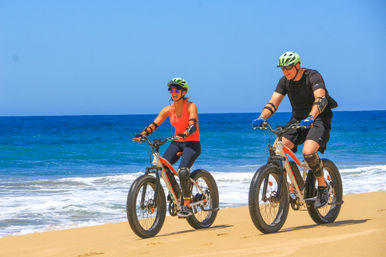Two cyclists wearing helmets and protective gear riding fat‑tire bikes along a sunny sandy beach with clear blue ocean waves and sky.
