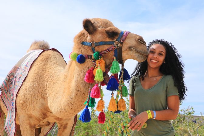 Smiling woman with curly hair poses outdoors holding a rope beside a tan camel decorated with colorful tassels and a patterned saddle blanket under a bright blue sky.