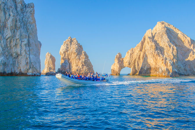Speedboat of passengers in life vests speeding past the iconic El Arco limestone rock formations at Land's End, Cabo San Lucas, Mexico, with sparkling blue water and a clear sky