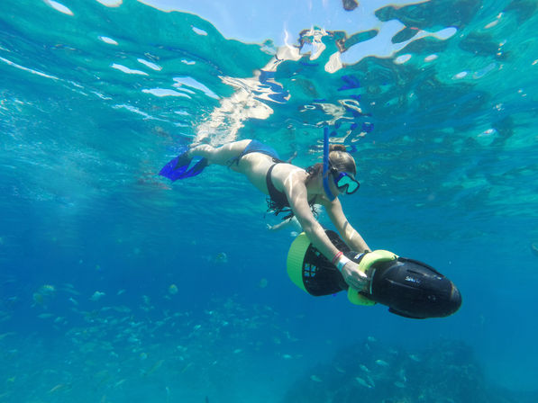 Snorkeler gliding with an underwater sea scooter in clear turquoise tropical ocean above a reef dotted with schooling fish