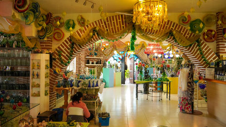 Playful, colorful artisan glass gallery inside red-brick arches — chandeliers and handblown glass vases and sculptures fill tables and shelves, with a person seated arranging pieces in the bright boutique.