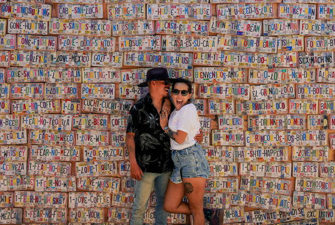Happy couple hugging and laughing in front of a vibrant wall of colorful novelty license-plate signs with playful travel and drink phrases — fun street-art vacation photo.