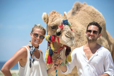 Smiling couple in sunglasses posing with a camel adorned with colorful tassels on a sunny beach