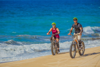 Two cyclists wearing neon-green helmets and knee pads ride fat-tire bikes on a sunny sandy beach beside turquoise ocean waves.