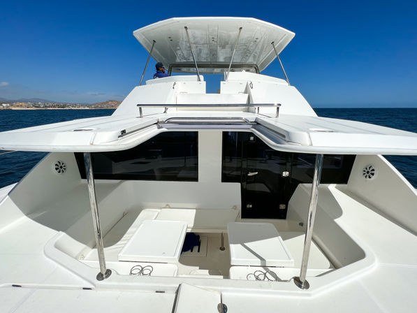 Sun-drenched front deck of a white luxury yacht with shaded flybridge, stainless steel supports, cushioned benches and folding tables, calm blue ocean and distant coastline under a clear sky.